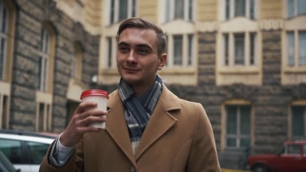 Молодой бизнесмен в плаще идет по улице в облачную погоду и пьет кофе. Медленное движение — стоковое видео