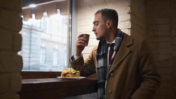 Mladý muž sedí v The Cafe a pití kávy z papíru Cup.The chlap je oblečen do pláště — Stock video