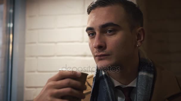 Молодой человек сидит в кафе и пьет кофе из бумажного кубка. Парень одет в плащ — стоковое видео