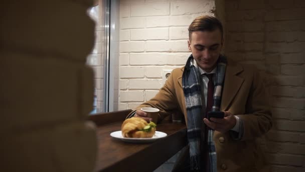 Ung Man sitter i The Cafe, med smartphone och dricka fika från ett papper Cup.The killen är klädd i mantel — Stockvideo