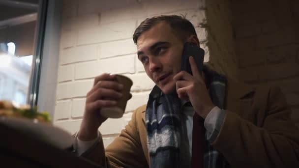 Ung Man sitter i The Cafe nära fönstret, dricka fika från ett papper kopp och skeaks via telefon. Killen är klädd i mantel — Stockvideo