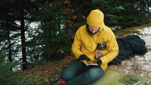 Ένας νεαρός άνδρας κάθεται κάτω από το λόφο του βουνού δίπλα από τα καταπράσινα δέντρα και ψάχνει για ένα νέο δρόμο. Κρατάει ένα δισκίο, ένα χάρτη, μια φωτογραφική μηχανή και ένα θερμός — Αρχείο Βίντεο