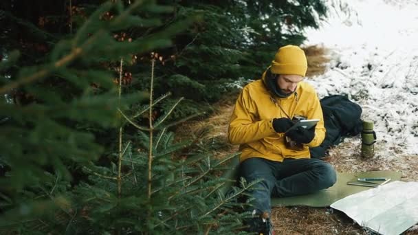 Сидя на горном холме рядом с зелеными деревьями, молодой человек пользуется табличкой — стоковое видео