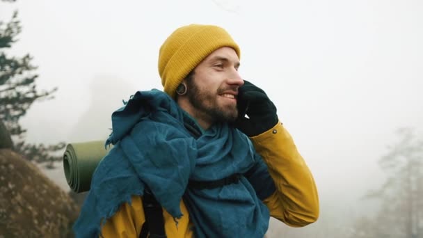 Man with beard, wearing yellow winter clothes talking on the phone. A hiker goes in the winter mountains with a backpack. Beautiful mountains in winter time — Stock Video