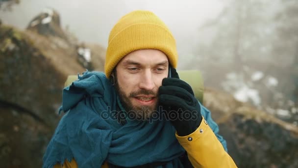 男人带着胡子, 穿着黄色的冬衣在电话里说话。一个徒步旅行者带着背包去冬天的山里。美丽的山在冬天时间 — 图库视频影像