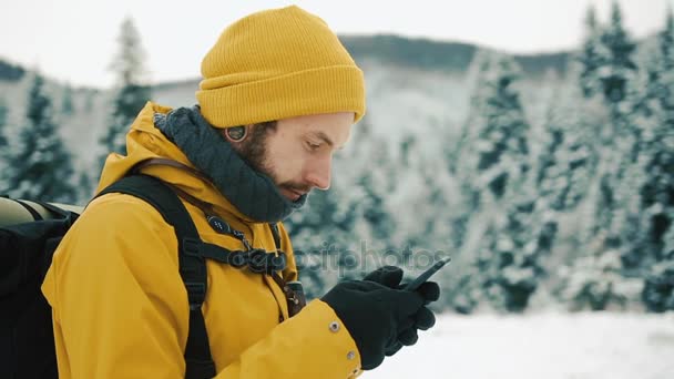 Uomo con la barba, indossa vestiti gialli invernali usando un telefono. Un escursionista va sulle montagne invernali con uno zaino. Belle montagne in inverno — Video Stock