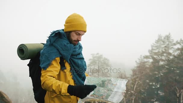 Férfi szakállal, sárga téli ruhában tartja a Térkép, ellenőrzése módon. Hegyek, téli időben, a háttérben — Stock videók