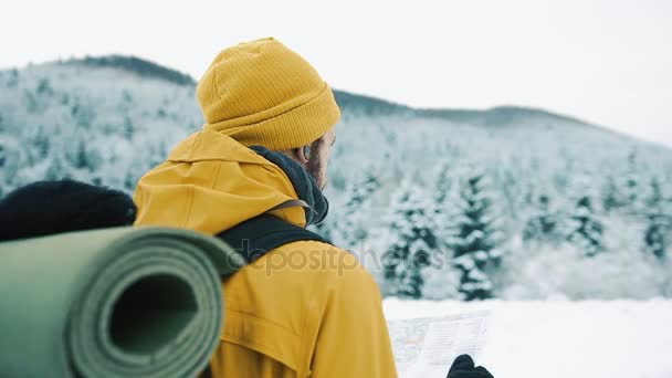 Lindas montanhas no inverno. Um cara com barba, vestindo roupas amarelas de inverno está vagando e segurando um mapa em suas mãos, contra o fundo da paisagem de inverno — Vídeo de Stock