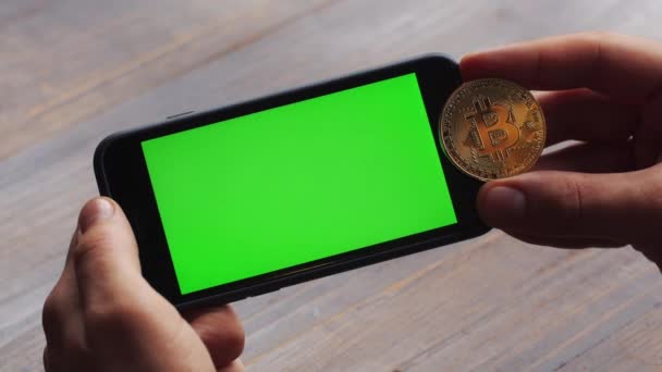 Homme utilisant un smartphone horizontal avec écran vert. Gros plan des mains mans avec téléphone mobile et une pièce de monnaie Bitcoin or. Clé chromatique. Ferme là. Horizontal — Video