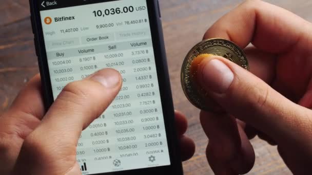 纽约, 美国-2018年1月28日: 一名男子在手机上使用 Bitfinex 网站。男人用比特币和手机, 双手紧紧的。比特币汇率兑换为美元图表。比特币价格 — 图库视频影像