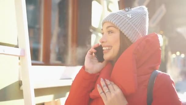 Piros téli kabát a gyönyörű nő áll az utcán, fedett hóban és beszél a smartphone a fényes nap — Stock videók