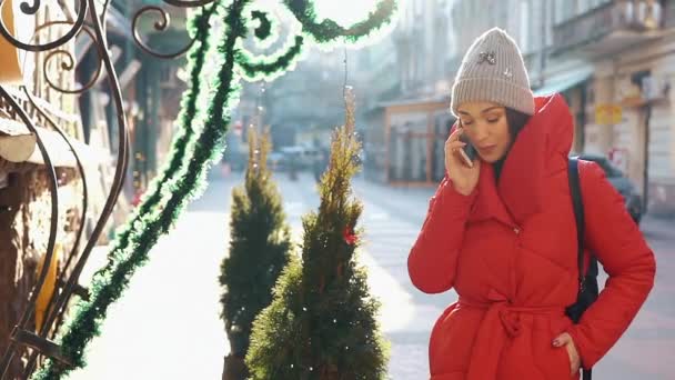 Hermosa mujer en chaqueta de invierno rojo se encuentra en la calle cubierta de nieve y habla en el teléfono inteligente en un día brillante — Vídeo de stock