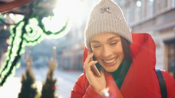 Mooie vrouw in rode winter jas staat op de straat die bedekt met sneeuw en gesprekken op de smartphone in een stralende dag — Stockvideo