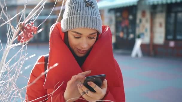 Ragazza in giacca invernale rossa digita qualcosa nel suo telefono in piedi sulla strada invernale — Video Stock