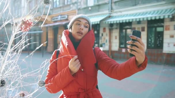 Charmante meisje in de rode winter jas neemt selfie op haar telefoon staande op de straat in een stralende dag winter — Stockvideo