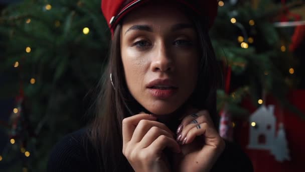 Modelo encantador posa en sombrero rojo de invierno ante un árbol de Navidad — Vídeos de Stock