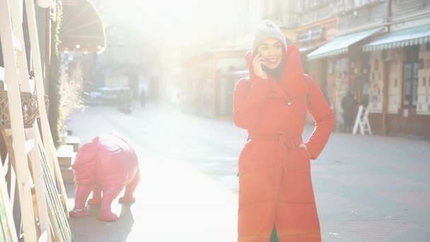 Hermosa mujer en chaqueta de invierno rojo se encuentra en la calle cubierta de nieve y habla en el teléfono inteligente en un día brillante — Vídeo de stock