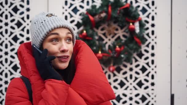 Красивая девушка в красной куртке разговаривает по смартфону, стоящему на улице в яркий зимний день — стоковое видео