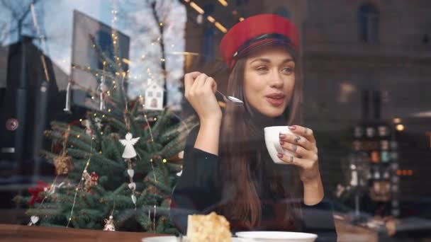 Look from the outside at charming woman in red hat drinking coffee sitting in the cosy cafe — Stock Video