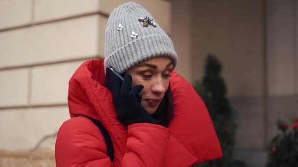 Красивая девушка в красной куртке разговаривает по смартфону, стоящему на улице в яркий зимний день — стоковое видео