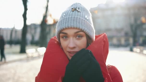 Portrét nádherné dívky v šedý klobouk a červený plášť s úsměvem, zatímco ona stojí na ulici před vánoční ozdoby v slunný zimní den — Stock video