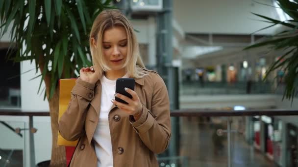 Chica comprueba su teléfono de pie con bolsas de compras en su hombro en el centro comercial — Vídeos de Stock