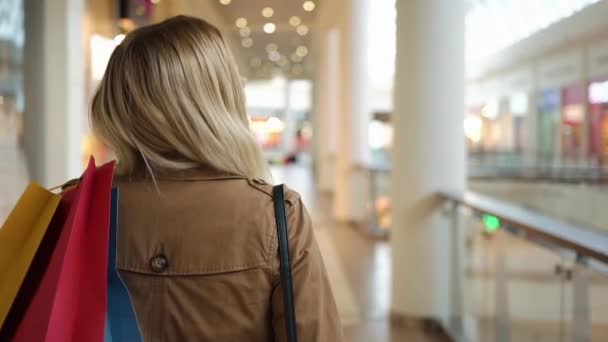 Szczęśliwy, blond kobieta uśmiecha się i patrzy na jej ramieniu, chodzenie z torby na zakupy wokół centrum handlowego — Wideo stockowe
