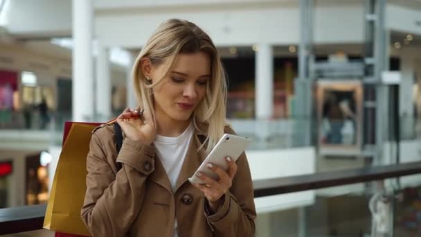 Meisje controleert haar telefoon staan met boodschappentassen op haar schouder in het winkelcentrum — Stockvideo