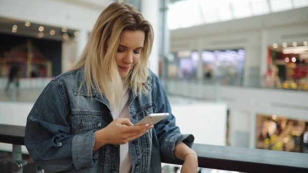 Молода жінка посміхається і друкує щось у своєму смартфоні, стоячи з сумками в торговому центрі — стокове відео