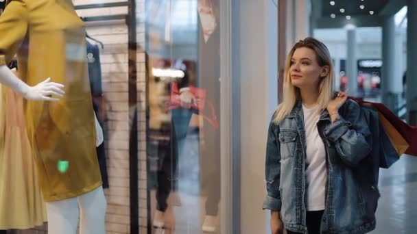 Encantada jovem loira caminha ao longo de uma janela de show com sacos no shopping — Vídeo de Stock