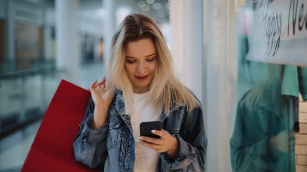 Sonriente chica rubia lee algo en su teléfono caminando con bolsas de compras alrededor de un centro comercial — Vídeo de stock