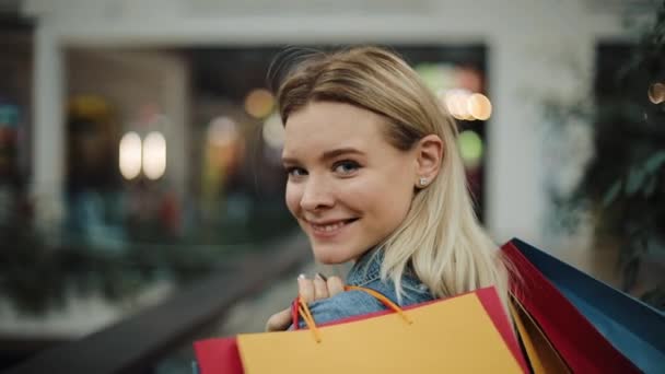 Blond meisje kijkt over haar schouder lopen met boodschappentassen in het winkelcentrum — Stockvideo