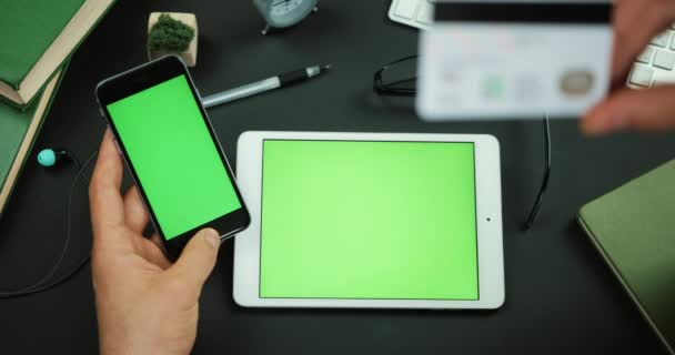 Mann hält Smartphone mit grünem Bildschirm und tippt Informationen von Kreditkarte auf einen Tisch mit grünem Bildschirm — Stockvideo