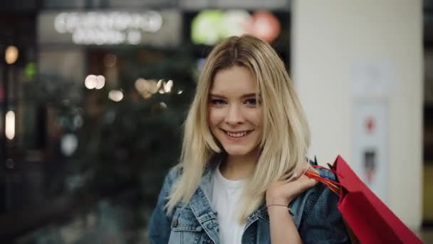 Молода блондинка в джинсовій куртці гуляє по торговому центру з різнокольоровими сумками — стокове відео