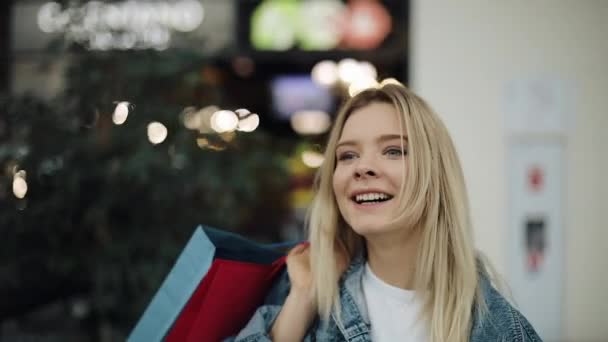 Jonge blonde vrouw in jeans jasje loopt rond een winkelcentrum met kleurrijke tassen — Stockvideo