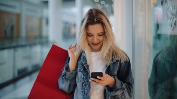 Souriant blonde fille types quelque chose dans son téléphone marche avec des sacs à provisions autour d'un centre commercial — Video