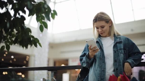 Donna bionda sta con le borse della spesa nel centro commerciale e digita qualcosa nel suo smartphone — Video Stock