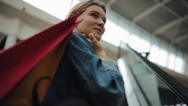 Unga blonda kvinnan i jeans jacka leenden gå upp på den rörliga trappor i köpcentrum — Stockvideo