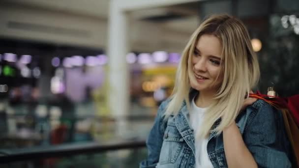 Giovane donna bionda in giacca di jeans cammina intorno a un centro commerciale con borse colorate — Video Stock