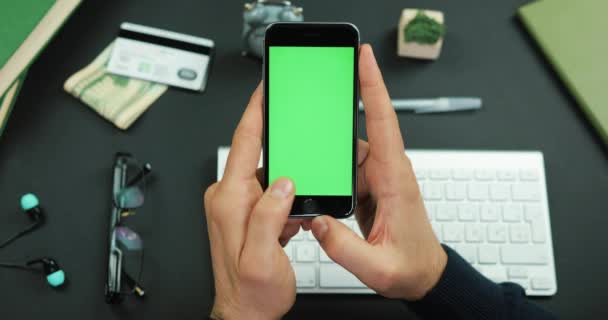 Ο άνθρωπος κατέχει ένα μαύρο smartphone με πράσινη οθόνη πάνω από ένα τραπέζι εργασίας και ειλητάρια κάτι για αυτό — Αρχείο Βίντεο