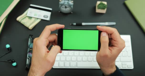Mannen innehar en svart smartphone med grön skärm över ett arbetsbord och kranar något på det — Stockvideo