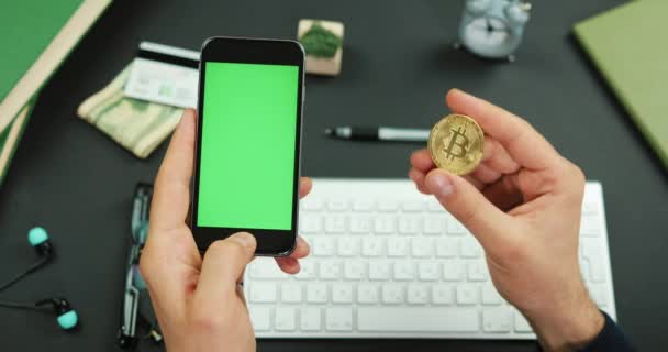 El hombre sostiene un teléfono inteligente negro con pantalla verde sobre una mesa de trabajo y un Bitcoin dorado — Vídeo de stock