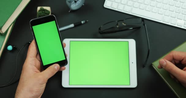 El hombre sostiene el teléfono inteligente con pantalla verde sobre una mesa con pantalla verde y toca algo en ella — Vídeo de stock