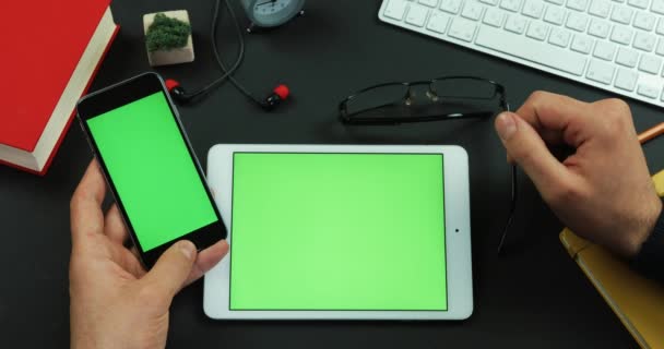 El hombre sostiene el teléfono inteligente con pantalla verde sobre una mesa con pantalla verde y toca algo en ella — Vídeo de stock