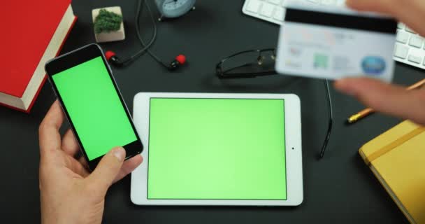 El hombre tiene un teléfono inteligente con pantalla verde y escribe información de la tarjeta de crédito en una mesa con pantalla verde — Vídeo de stock