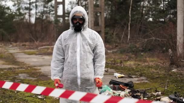 Людина в біо-небезпечному костюмі і газових масках стоїть за смугою на території, повній відходів — стокове відео