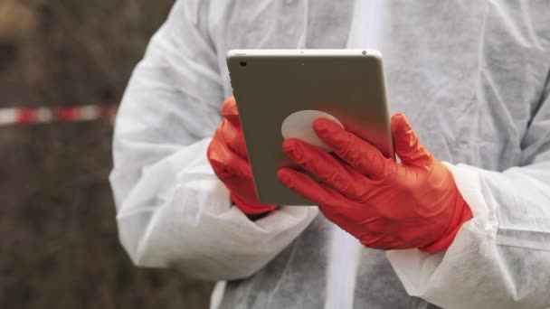 Hombre en traje de bio-peligro y máscara de gas toma notas en su tableta de pie en la tierra contaminada — Vídeo de stock
