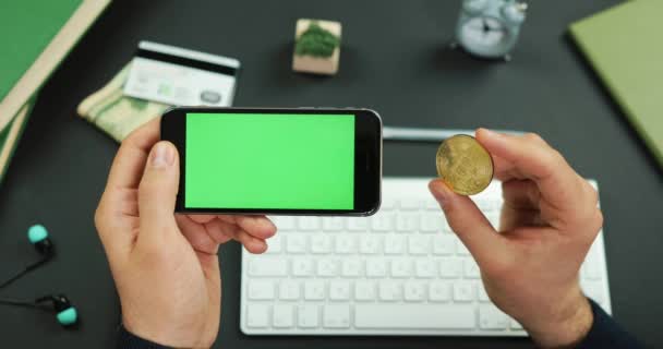 Ο άνθρωπος κατέχει ένα μαύρο smartphone με πράσινη οθόνη πάνω από έναν λειτουργώντας πίνακα και μια χρυσή Bitcoin — Αρχείο Βίντεο