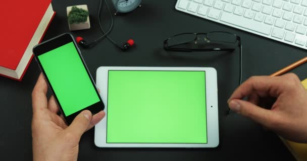 El hombre sostiene el teléfono inteligente con pantalla verde sobre una mesa con pantalla verde y toca algo en ella — Vídeo de stock