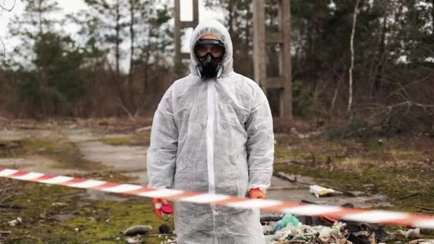 Człowiek w zagrożenia biologicznego garnitur i maski gazowe stoi za paskiem na terytorium pełen odpadów — Wideo stockowe